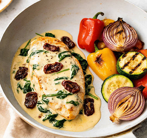 Creamy Tuscan chicken with sun-dried tomatoes and spinach in a skillet, served with grilled bell peppers and zucchini