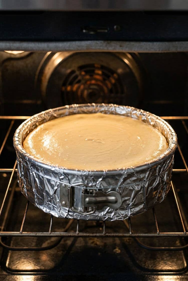 Whole New York cheesecake baking to a pale golden finish inside a home oven on the center wire rack in a double foil-wrapped springform pan