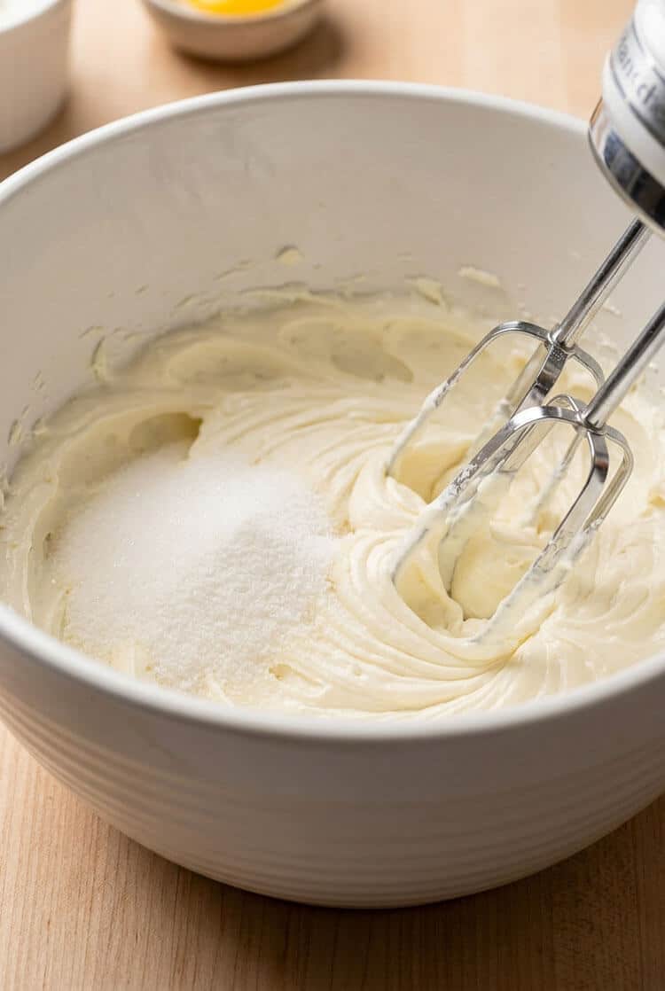 Electric hand mixer beating softened room temperature block cream cheese and granulated sugar into a smooth, glossy cream in a white ceramic mixing bowl