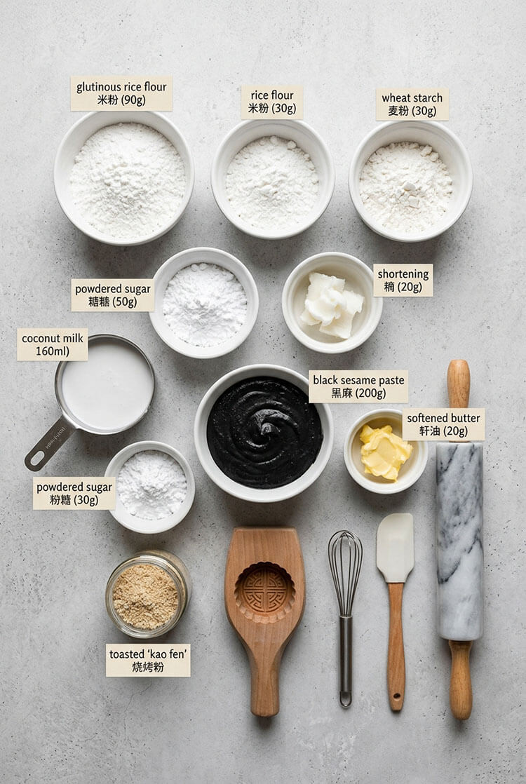 Flatlay arrangement of white bowls filled with glutinous rice flour, rice flour, wheat starch, powdered sugar, shortening, coconut milk, roasted black sesame paste, butter, toasted kao fen, wooden mold, and rolling pin on a grey background