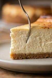 Silver dessert fork sinking into a tall, ultra-dense slice of creamy New York style cheesecake showing a thick, velvety crumb and golden graham crust