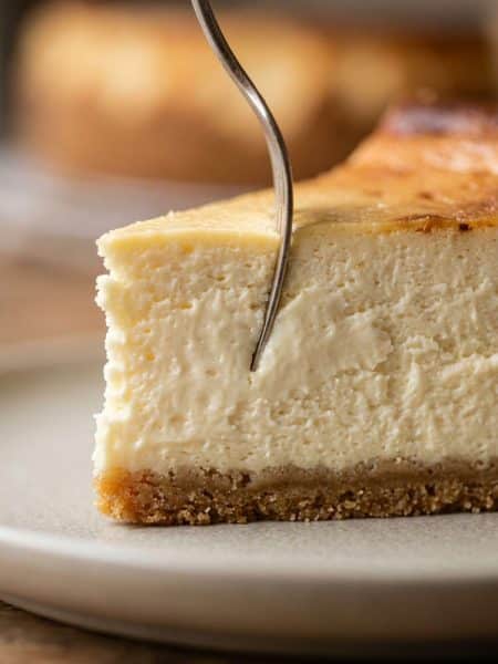 Silver dessert fork sinking into a tall, ultra-dense slice of creamy New York style cheesecake showing a thick, velvety crumb and golden graham crust