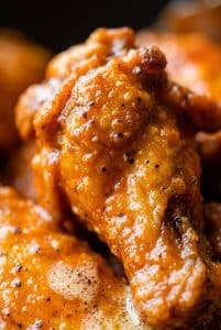 Macro extreme close-up of a glossy, fiery hot buffalo chicken drumette showing blistered crispy skin coated in red-orange pepper sauce and cracked black pepper