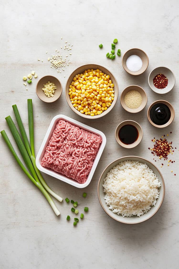 Flatlay arrangement of raw ground pork, sweet yellow corn kernels, steamed white rice, green scallions, minced garlic, soy sauce, and chili flakes on a light marble surface