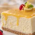 Macro close-up of a slice of creamy no-bake lemon cheesecake with glossy golden lemon curd dripping down the side, garnished with a fresh raspberry and lemon peel