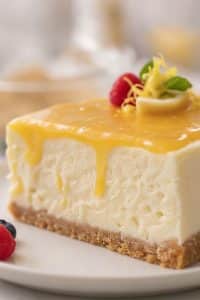 Macro close-up of a slice of creamy no-bake lemon cheesecake with glossy golden lemon curd dripping down the side, garnished with a fresh raspberry and lemon peel