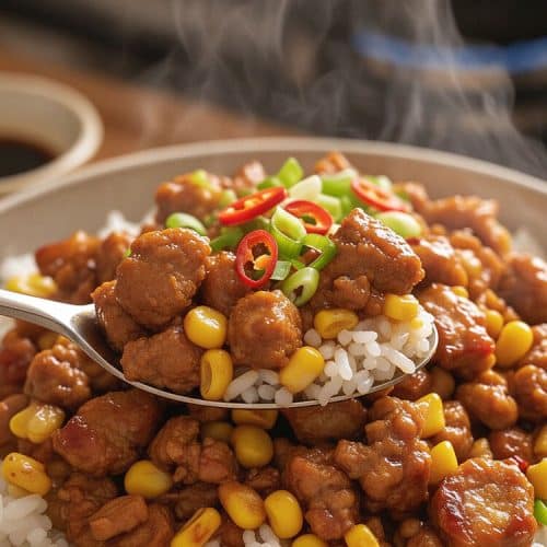 A metal spoon lifting a steaming bite of glossy ground pork, sweet corn, and fluffy white jasmine rice garnished with scallions and red chili slices