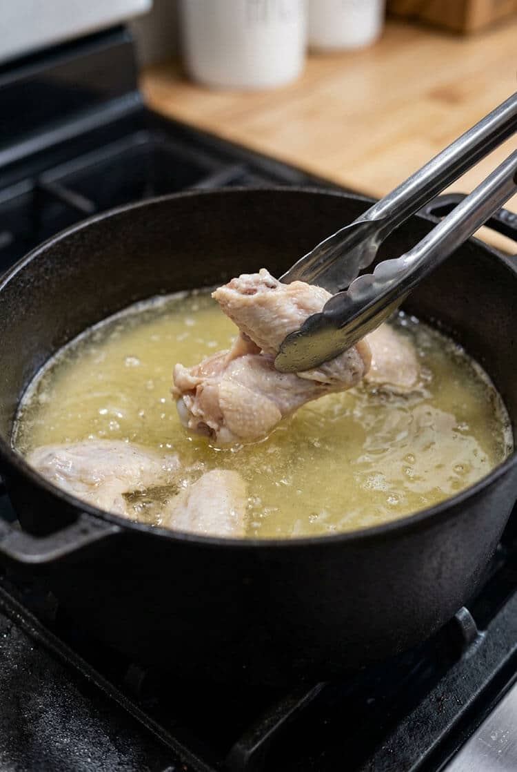 Metal tongs lifting a pale, lightly cooked chicken wing out of a bubbling cast iron pot of 250 degree frying oil