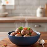 Steaming hot bouncy beef balls piled in a blue bowl garnished with sliced red chili, scallions, and cilantro on a wooden board