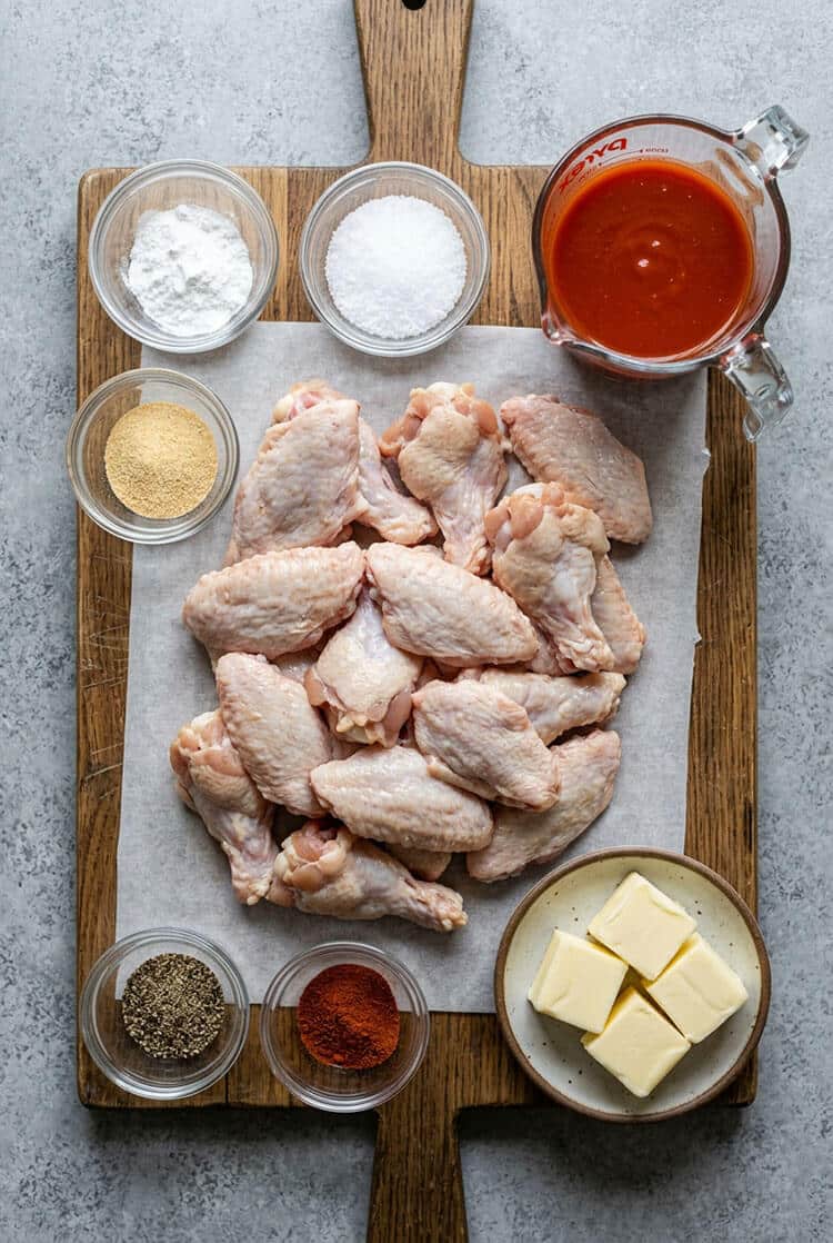 Flatlay arrangement of fresh split chicken wings on parchment paper, Frank’s RedHot hot sauce in a glass measuring cup, cubed cold butter, baking powder, kosher salt, garlic powder, black pepper, and cayenne pepper on a wooden board