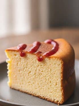 A tall, cotton-soft slice of Japanese soufflé cheesecake on a dark plate showing an ultra-fine crumb structure drizzled with glossy red strawberry coulis