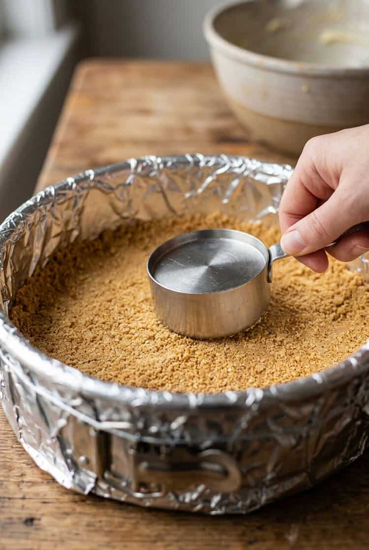 Hand firmly pressing buttery graham cracker crumbs into the bottom of a foil-wrapped 9-inch springform pan using the flat bottom of a stainless steel measuring cup