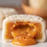 Close-up cross section of a warm white snowskin mooncake with steaming, glossy golden salted egg lava custard flowing out onto a white ceramic plate