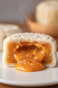 Close-up cross section of a warm white snowskin mooncake with steaming, glossy golden salted egg lava custard flowing out onto a white ceramic plate