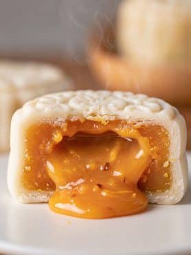 Close-up cross section of a warm white snowskin mooncake with steaming, glossy golden salted egg lava custard flowing out onto a white ceramic plate