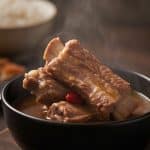 A hearty black bowl of steaming authentic Bak Kut Teh pork rib soup with tender meat on the bone, goji berries, and rich peppery broth on a wooden board