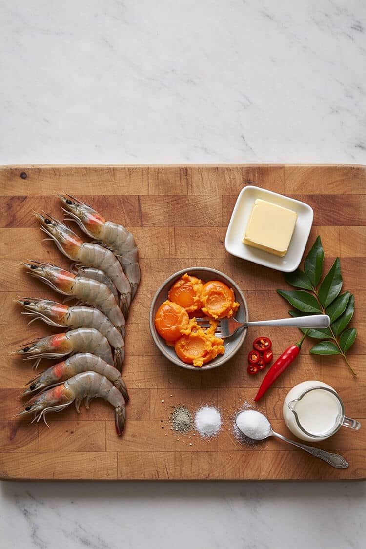 Overhead flatlay view of fresh whole prawns, mashed salted duck egg yolks in a bowl, butter block, fresh curry leaves, red chili, evaporated milk, cornstarch, salt, and white pepper on a wooden board