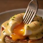 Macro close-up of a vintage silver fork piercing a poached egg covered in hollandaise sauce, with deep golden yolk oozing over a toasted English muffin