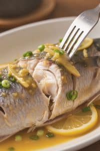 Flaking tender steamed mullet fish meat with a fork revealing moist white flakes coated in savory citrus sauce.