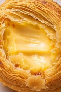 Close up cross section of a flaky egg tart showing paper-thin laminated pastry layers and silky custard center.