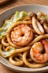 Macro close-up of glossy Japanese seafood udon noodles with charred shrimp, shimeji mushroom, and squid ring in a shallow bowl.