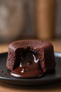 Macro close-up of an inverted molten chocolate lava cake sliced open on a black ceramic plate with a thick, glossy pool of dark chocolate flowing out
