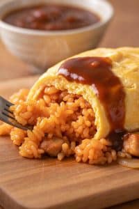 Macro close-up of a metal fork scooping a bite of caramelized ketchup fried rice with tender chicken from inside a soft, barely-set golden egg blanket coated in savory sauce
