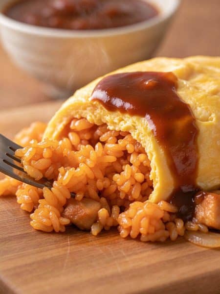 Macro close-up of a metal fork scooping a bite of caramelized ketchup fried rice with tender chicken from inside a soft, barely-set golden egg blanket coated in savory sauce