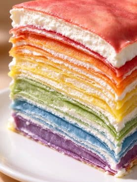 A slice of rainbow crepe cake recipe showing delicate colorful crepe layers filled with fluffy whipped cream.