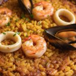 Macro close-up of tender seared shrimp, open black mussels, squid rings, and golden saffron Bomba rice with caramelized crispy socarrat edges