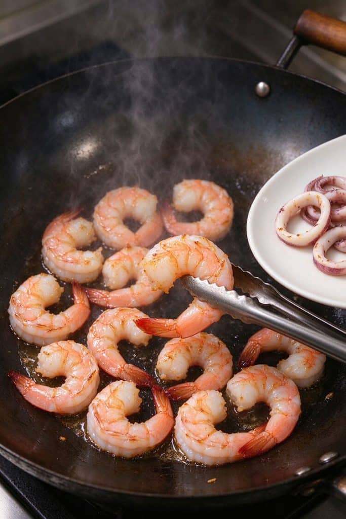 Flipping pink seared shrimp with metal tongs in a hot wok next to a small plate of sliced squid rings.