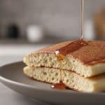 Macro close-up cross-section of a sliced fluffy pancake showing an airy, soft, cake-like interior crumb with a stream of golden maple syrup pouring over the golden top crust