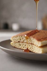 Macro close-up cross-section of a sliced fluffy pancake showing an airy, soft, cake-like interior crumb with a stream of golden maple syrup pouring over the golden top crust
