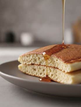 Macro close-up cross-section of a sliced fluffy pancake showing an airy, soft, cake-like interior crumb with a stream of golden maple syrup pouring over the golden top crust