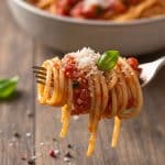 Macro close-up of a fork holding a neat twirl of glossy al dente spaghetti coated in thick tomato sauce, topped with grated Parmesan and a tiny fresh basil leaf