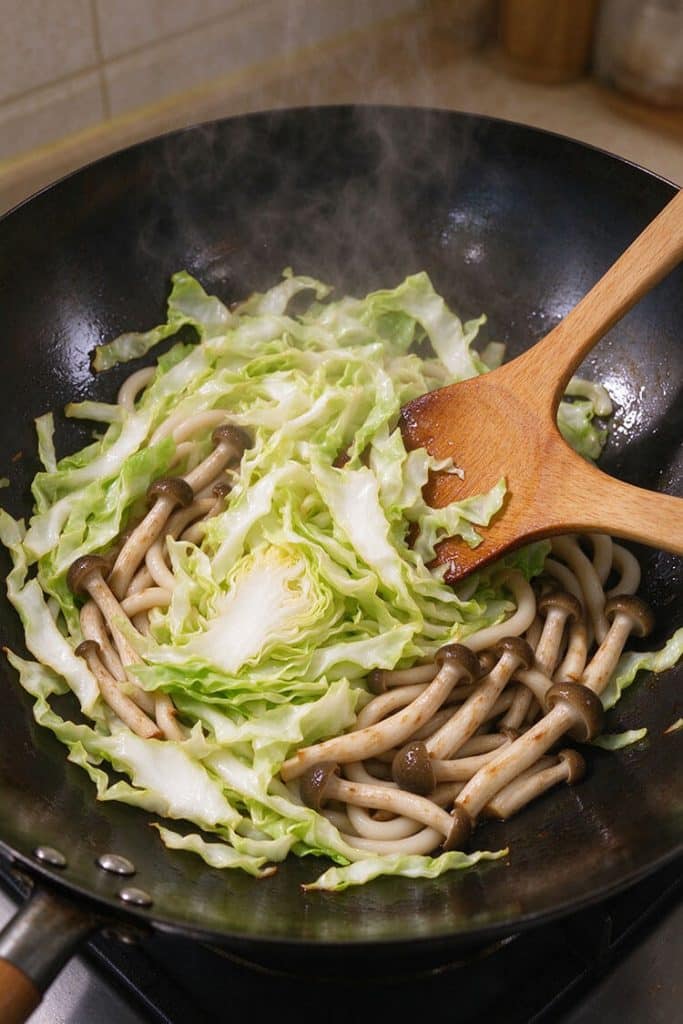 Stirring sliced green cabbage and shimeji mushrooms with a wooden spatula in a hot steaming black wok.