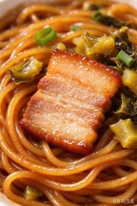 Extreme close up of a glossy tender pork belly slice resting over savory noodles and pickled greens.